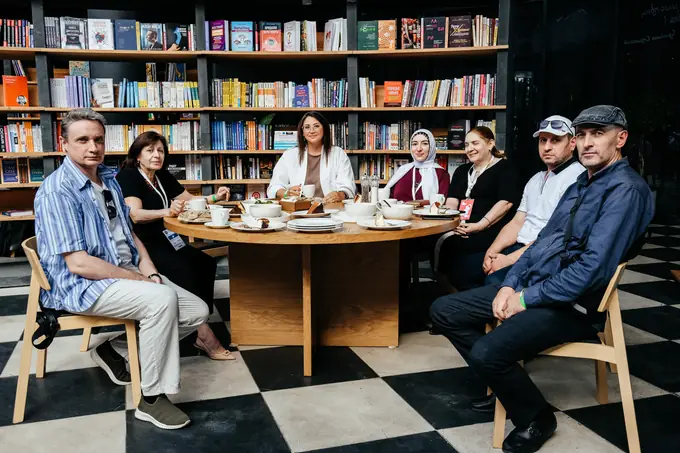 Гости за круглым столом с чаем и угощением на фоне книжных полок